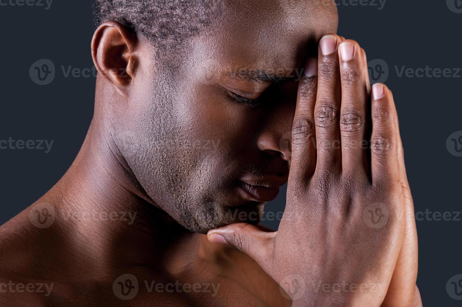 bidden Mens. kant visie van jong Afrikaanse Mens bidden terwijl Holding handen geklemd in de buurt en tegen grijs achtergrond foto