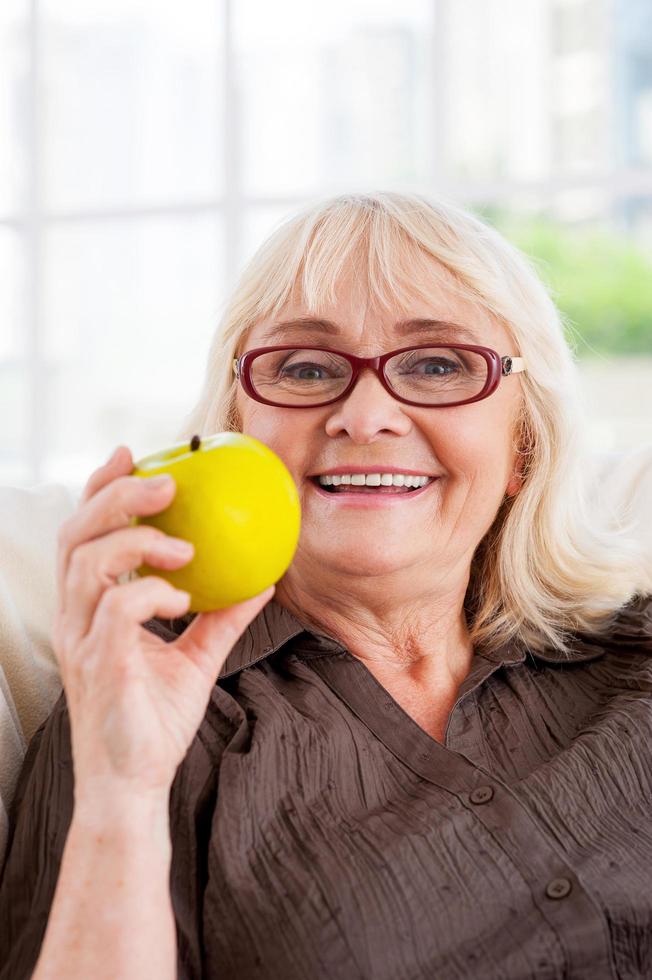 genieten van gezond levensstijl. vrolijk senior vrouw Holding appel en op zoek Bij camera terwijl zittend Bij de stoel foto