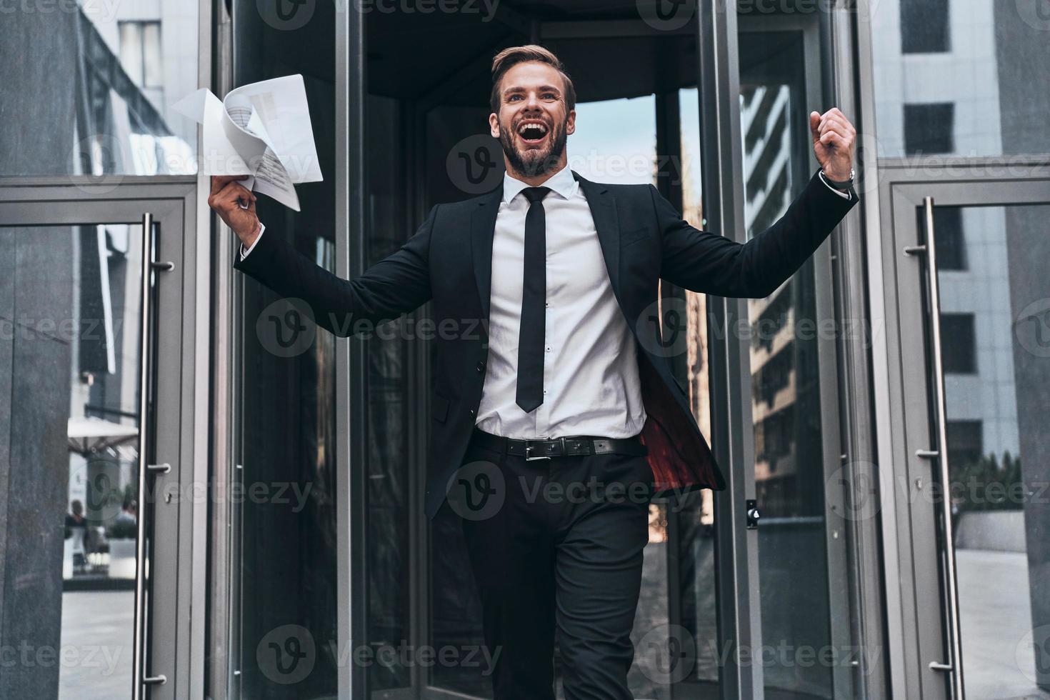 knap jong zakenman in vol pak glimlachen en Holding document terwijl wandelen buitenshuis foto