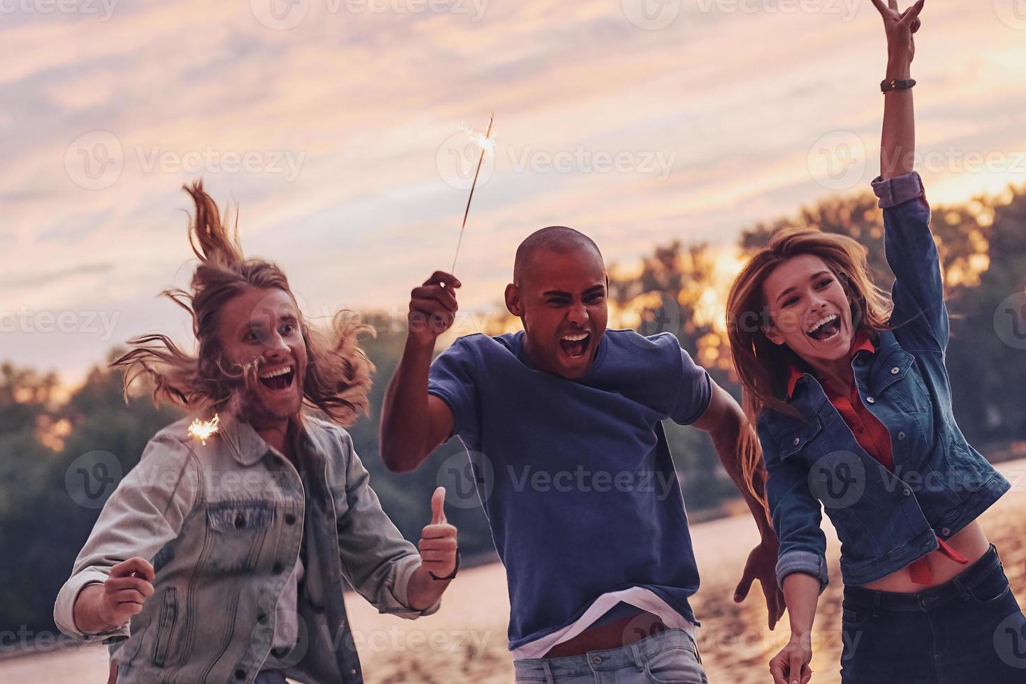 genieten van de moment. groep van jong mensen in gewoontjes slijtage glimlachen en gebaren terwijl jumping Aan de pier foto