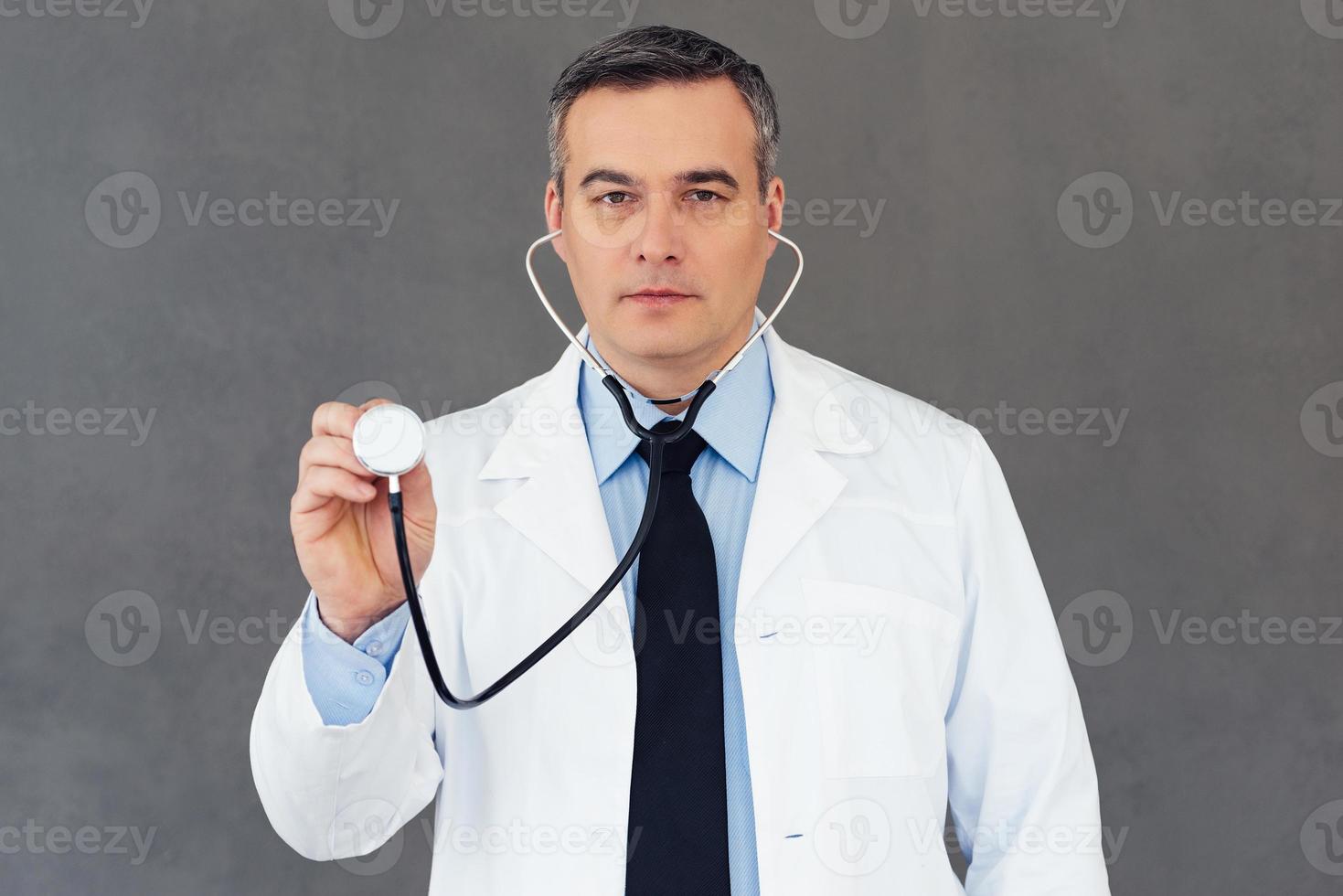 medisch examen. volwassen mannetje dokter Holding stethoscoop en op zoek Bij camera terwijl staand tegen grijs achtergrond foto