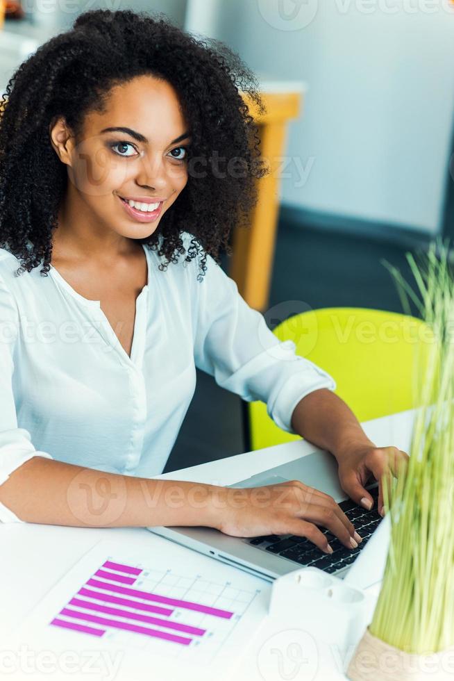 blijven positief Bij werk. glimlachen jong Afrikaanse Dames werken Aan laptop en op zoek Bij camera terwijl zittend Bij werken plaats foto