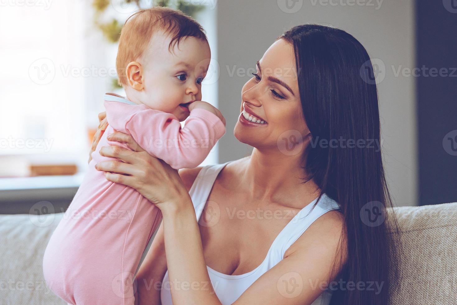 mijn zoet baby vrolijk mooi jong vrouw Holding baby meisje in haar handen en op zoek Bij haar met liefde terwijl zittend Aan de bankstel Bij huis foto