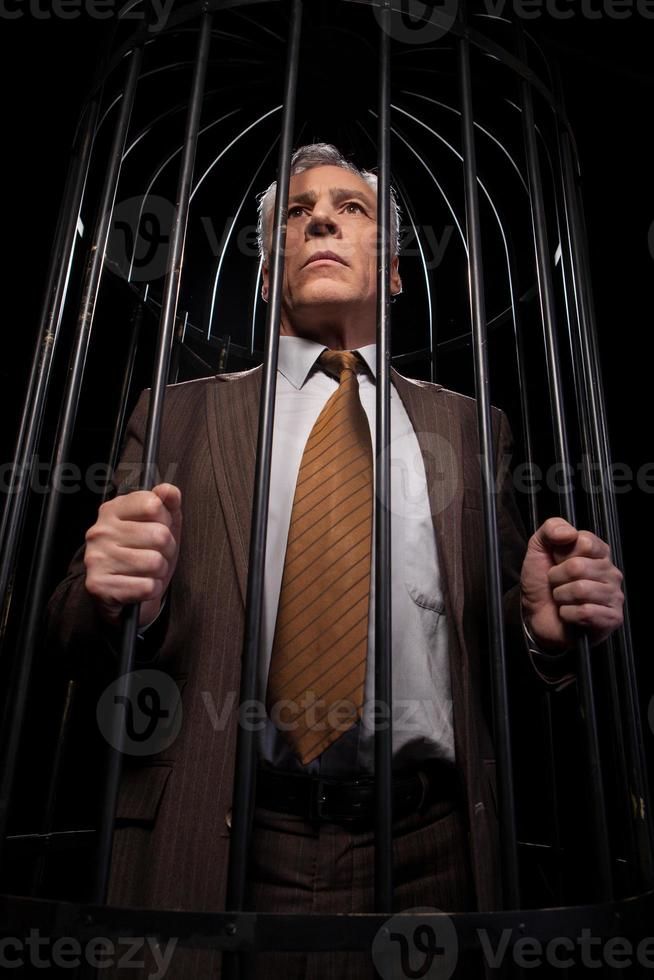 mannen in cel. laag hoek visie van geroest senior Mens in formele kleding staand in cel en op zoek weg terwijl geïsoleerd Aan zwart achtergrond foto