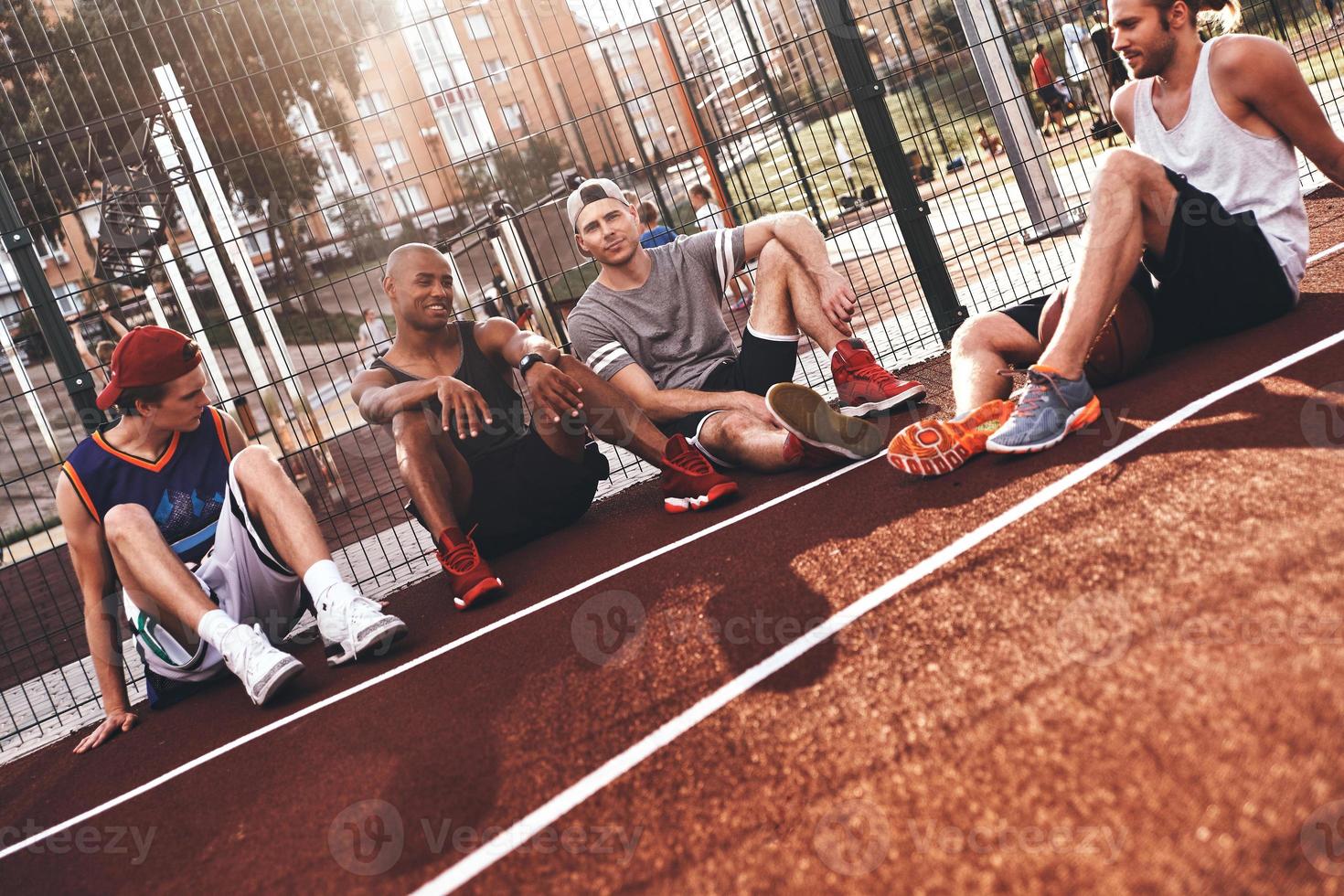 het beste vrienden. groep van jong mannen in sport- kleding glimlachen terwijl zittend Aan de basketbal veld- buitenshuis foto