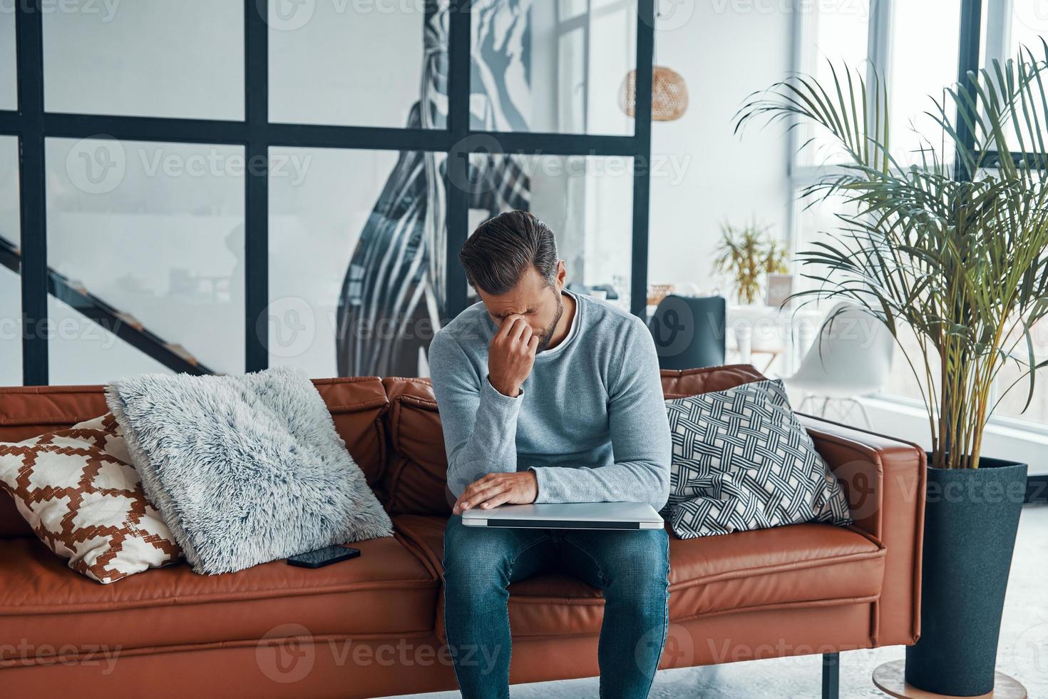 gefrustreerd jong Mens aanraken zijn hoofd en houden ogen Gesloten terwijl zittend Aan de sofa Bij huis foto