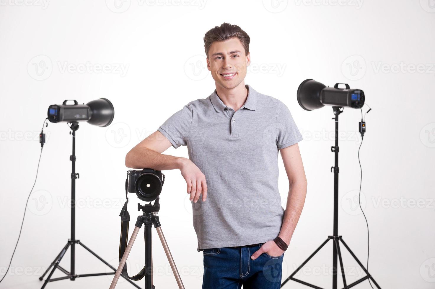 fotograaf in studio. knap jong Mens in polo overhemd leunend Bij de statief met camera terwijl staand in studio met verlichting uitrusting Aan achtergrond foto