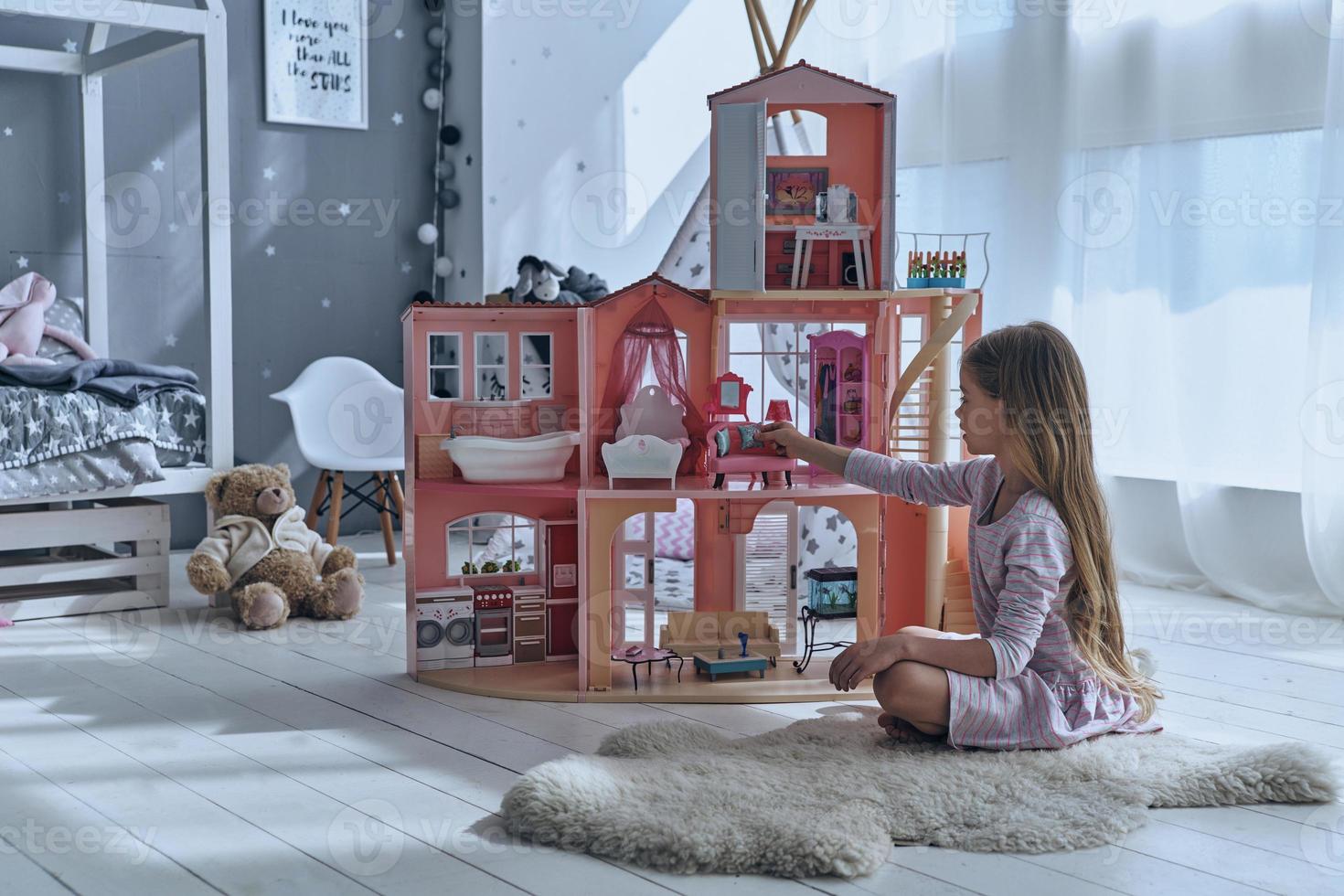 gevoel speels. schattig weinig meisje spelen met een poppenhuis terwijl zittend Aan de verdieping in slaapkamer foto