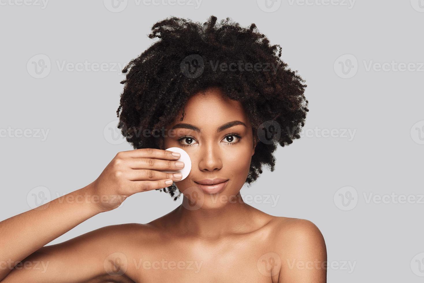verwennerij. aantrekkelijk jong Afrikaanse vrouw toepassen schoonmaak spons en glimlachen terwijl staand tegen grijs achtergrond foto