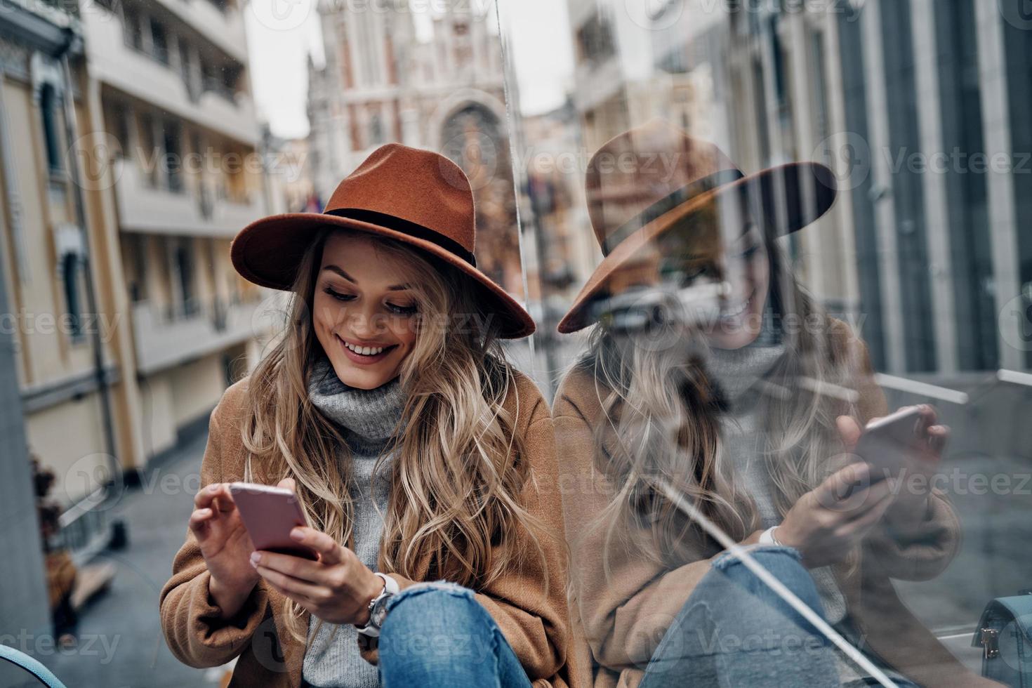 blijven verbonden. aantrekkelijk jong vrouw in hoed en jas gebruik makend van haar slim telefoon terwijl uitgeven zorgeloos tijd in de stad foto