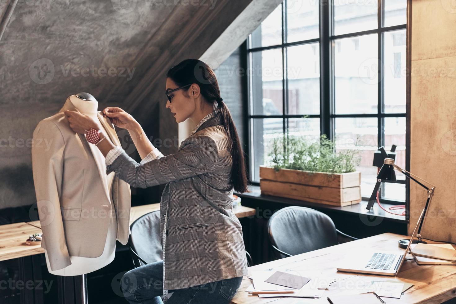 betalen aandacht naar elke detail. echt jong vrouw in eyewear aanpassen een halsband van de jasje Aan mannequin terwijl staand in haar werkplaats foto