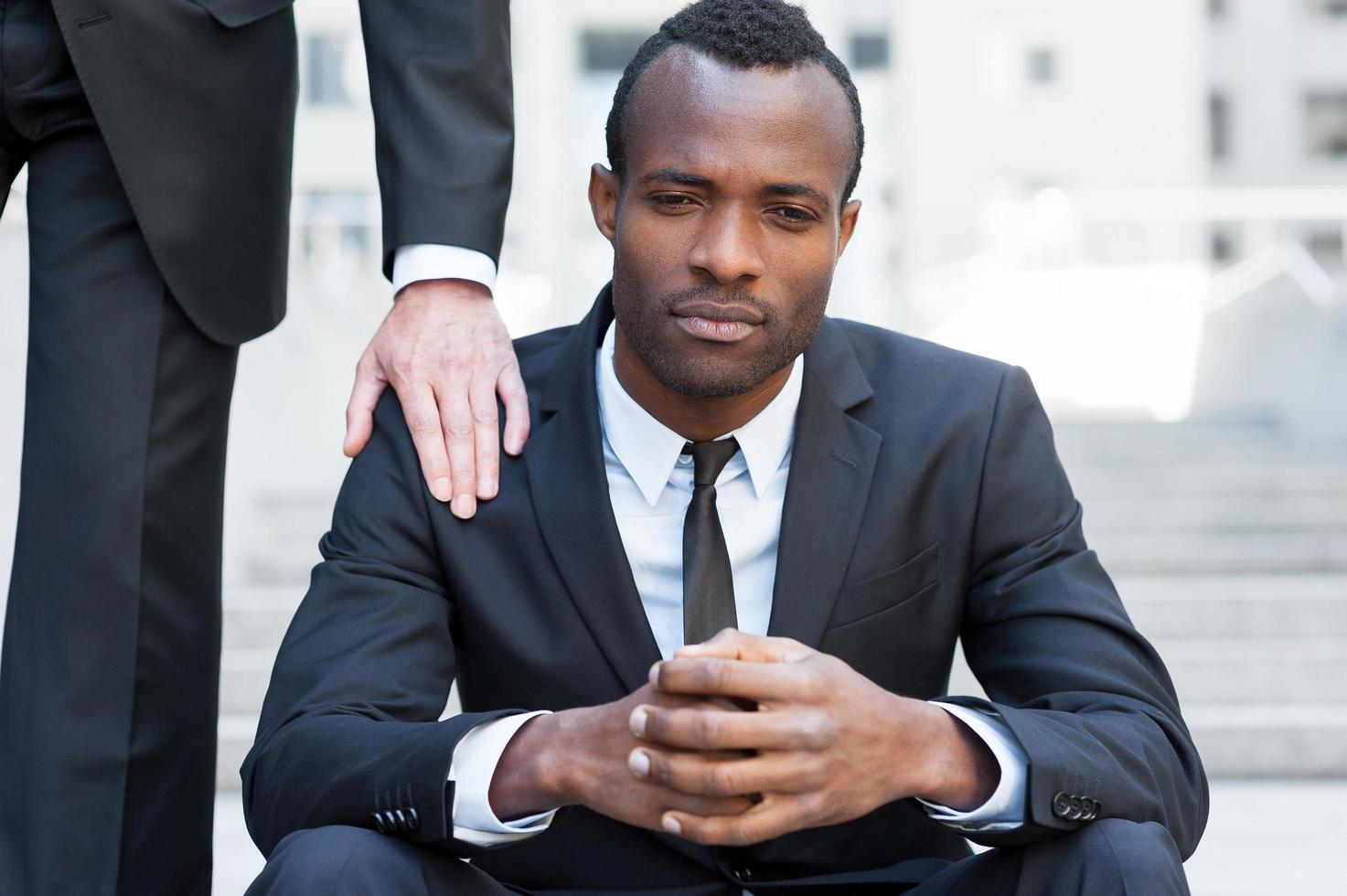 hij behoeften een vriendelijk steun. gefrustreerd jong Afrikaanse Mens in formele kleding zittend Aan trappenhuis terwijl iemand aanraken zijn schouder met hand- foto