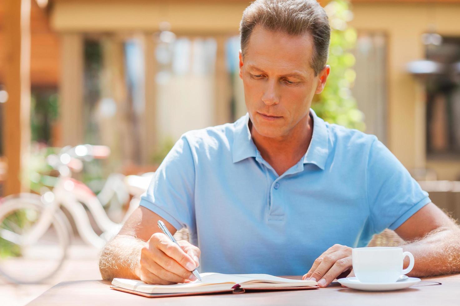 gebruik makend van moment naar maken sommige notities. zelfverzekerd volwassen Mens schrijven iets in zijn Notitie stootkussen terwijl zittend Bij de tafel buitenshuis met huis in de achtergrond foto