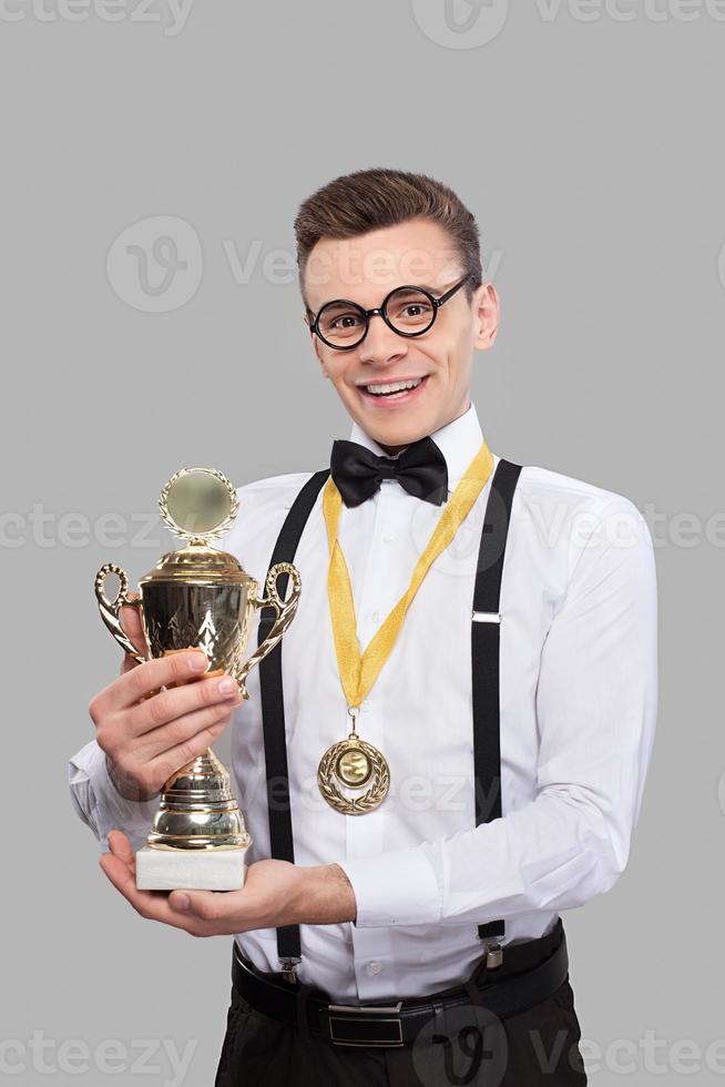 ik ben winnaar vrolijk jong Mens in boog stropdas Holding een trofee en glimlachen terwijl staand tegen grijs achtergrond foto