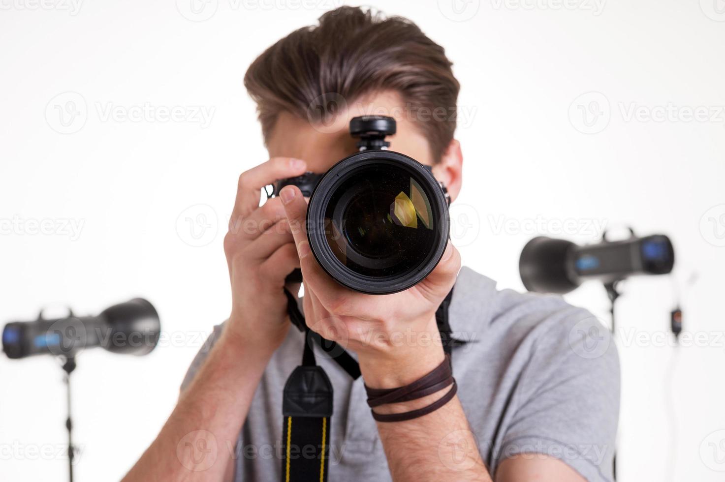 glimlach naar de camera jong Mens in polo overhemd het schieten u met digitaal camera terwijl staand in studio met verlichting uitrusting Aan achtergrond foto