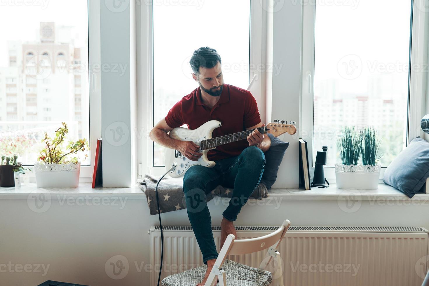 knap jong Mens in gewoontjes kleding spelen gitaar terwijl zittend Aan de venster dorpel foto