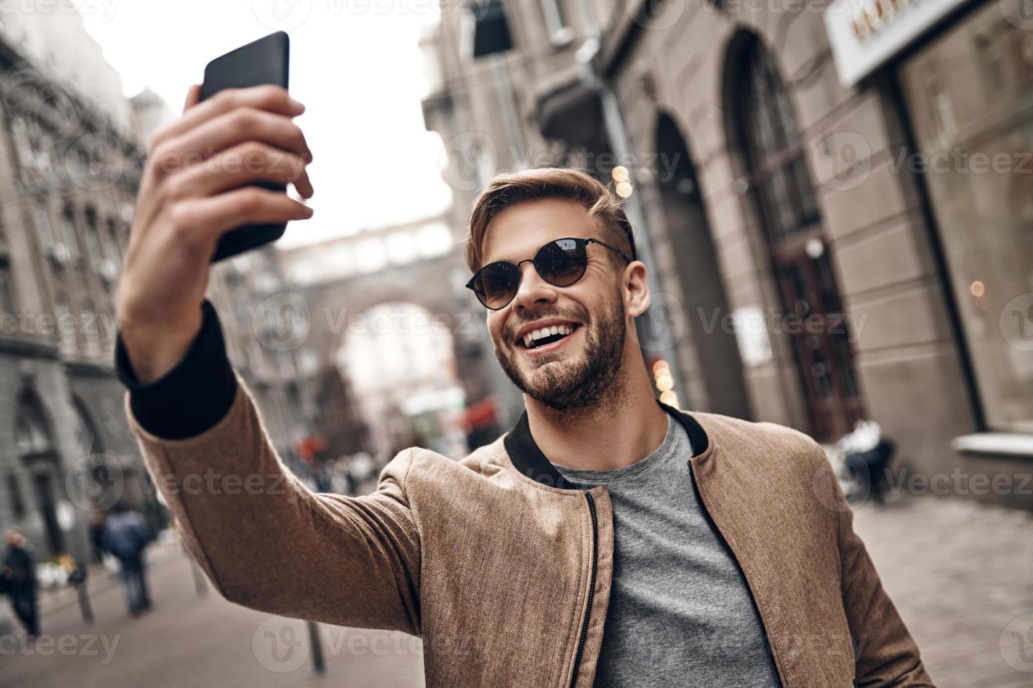 sharing gelukkig selfie. knap jong glimlachen Mens in gewoontjes slijtage nemen selfie gebruik makend van zijn slim telefoon terwijl staand buitenshuis foto