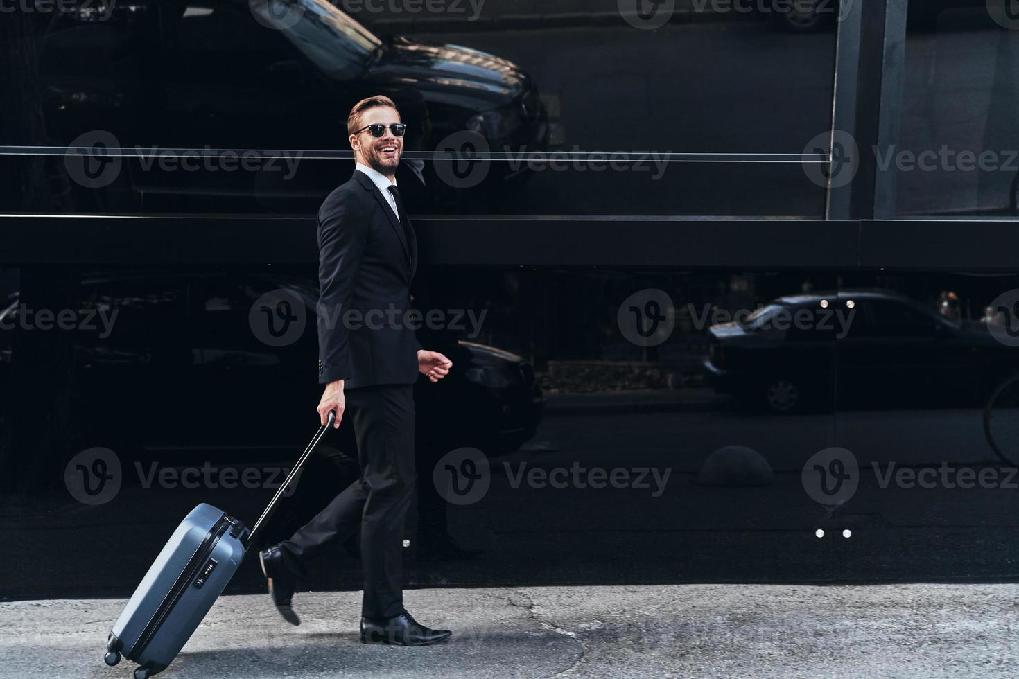 jong en succesvol. vol lengte van jong Mens in vol pak trekken bagage en glimlachen terwijl wandelen buitenshuis foto