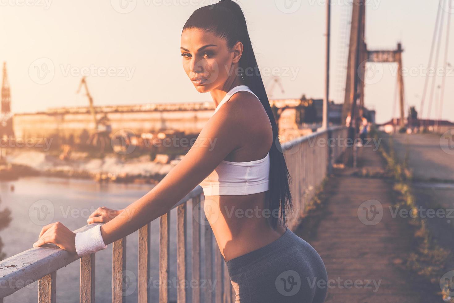 sportief vrouw. mooi jong vrouw in sport- kleding op zoek Bij camera terwijl staand Aan de brug met stedelijk visie in de achtergrond foto