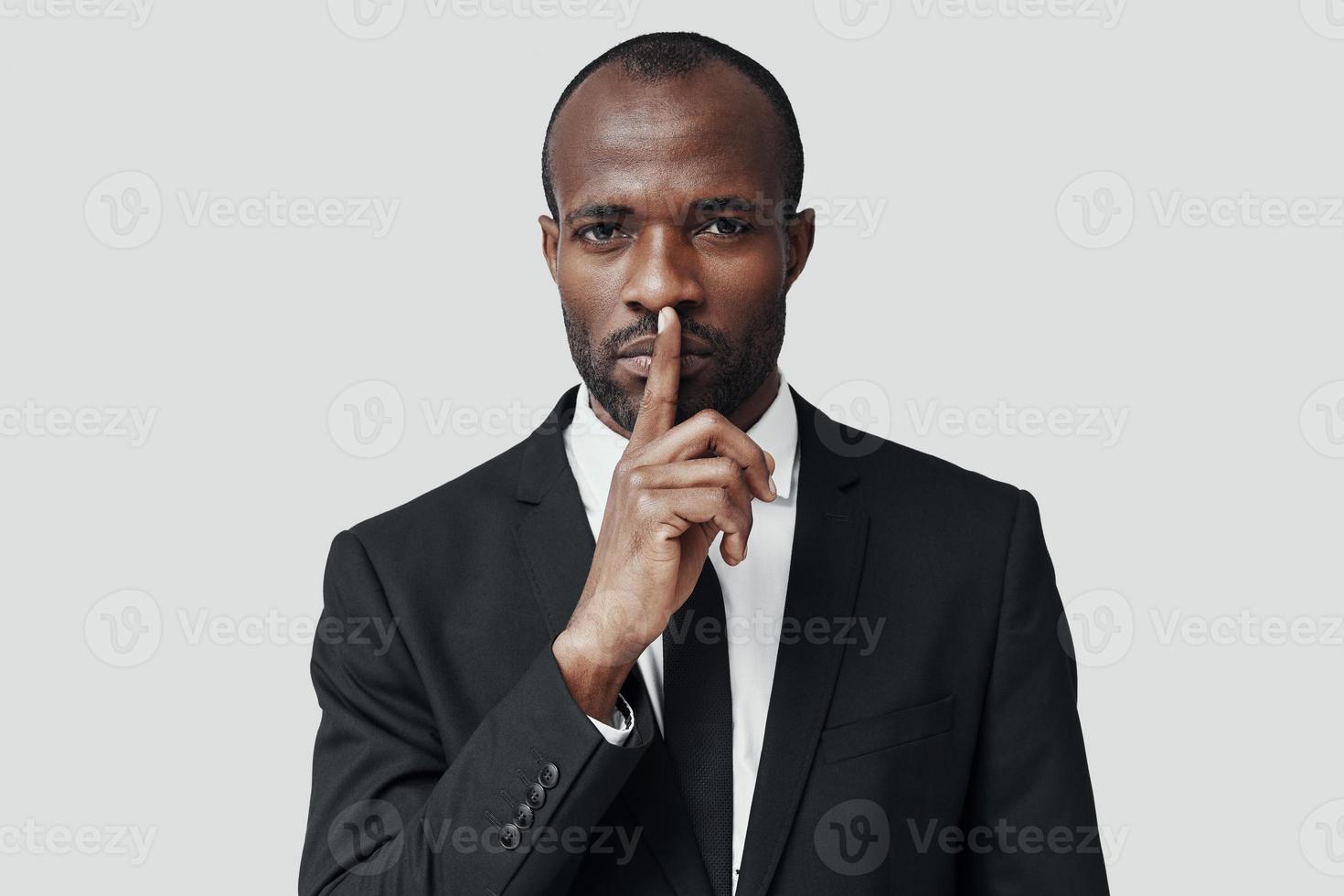 mysterieus jong Afrikaanse Mens in formele kleding houden vinger Aan lippen terwijl staand tegen grijs achtergrond foto