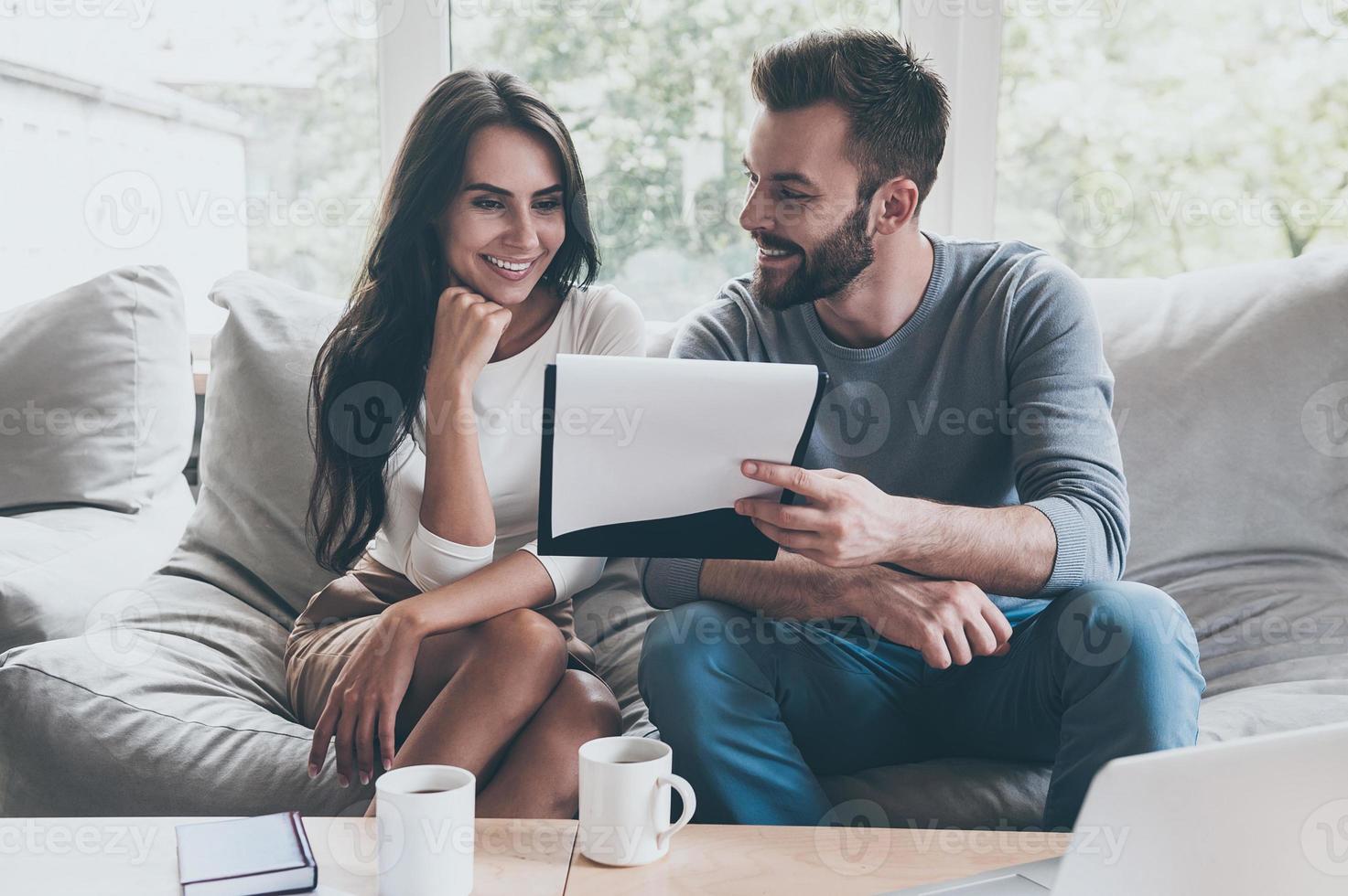 Super goed mogelijkheden voor ons vrolijk jong Mens tonen sommige documenten naar zijn vrouw zittend dichtbij naar hem Aan de bankstel foto