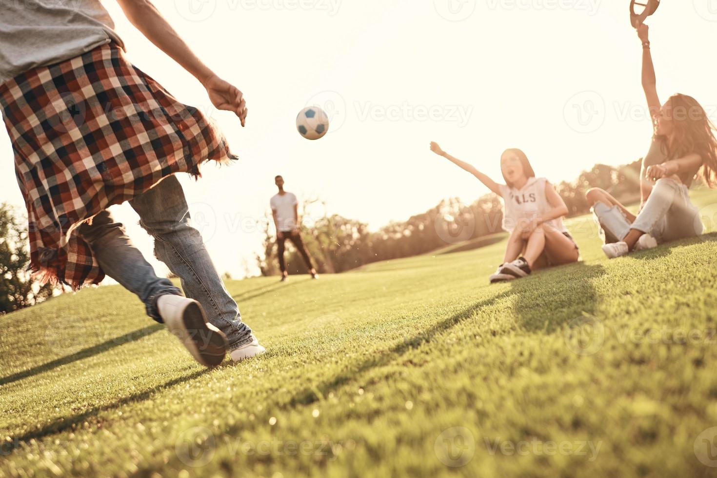 twee jong Dames zittend Aan de gras en gebaren terwijl hun mannetje vrienden spelen voetbal buitenshuis foto