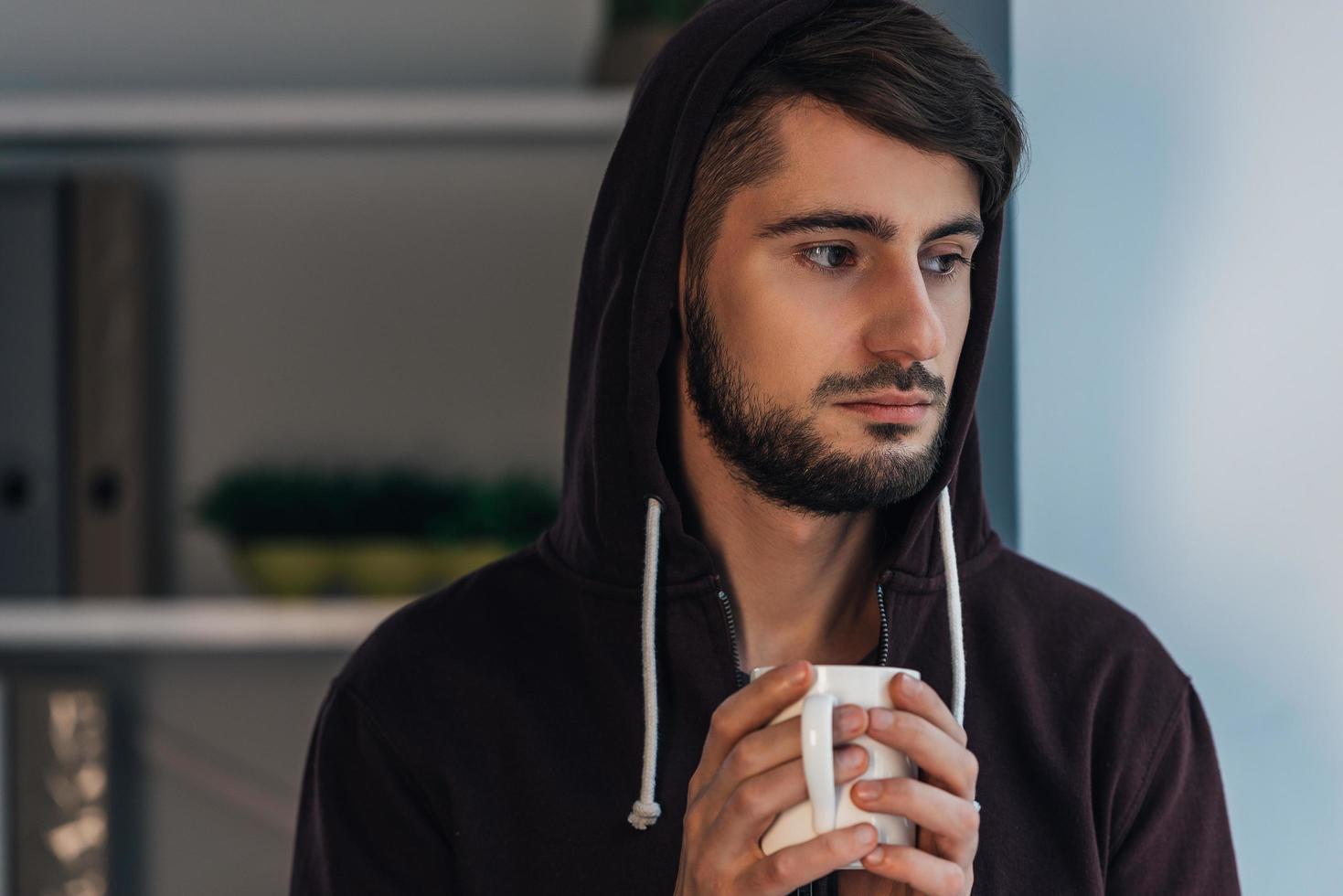 gevoel gefrustreerd. portret van nadenkend jong Mens in met een kap overhemd Holding koffie kop en op zoek weg terwijl staand Bij huis foto