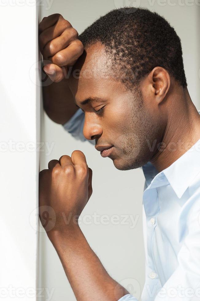 depressie en hopeloosheid. kant visie van depressief jong Afrikaanse Mens in blauw overhemd leunend Bij de muur en houden ogen Gesloten foto