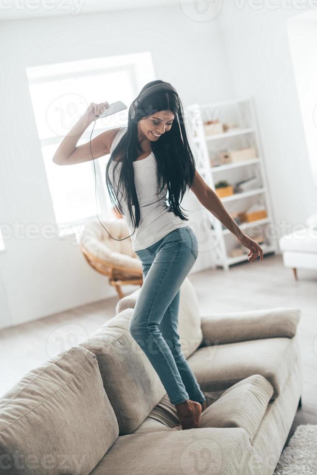 tijd naar dans vol lengte van aantrekkelijk jong vrouw in hoofdtelefoons Holding slim telefoon en gebaren terwijl dansen Aan de bankstel Bij huis foto