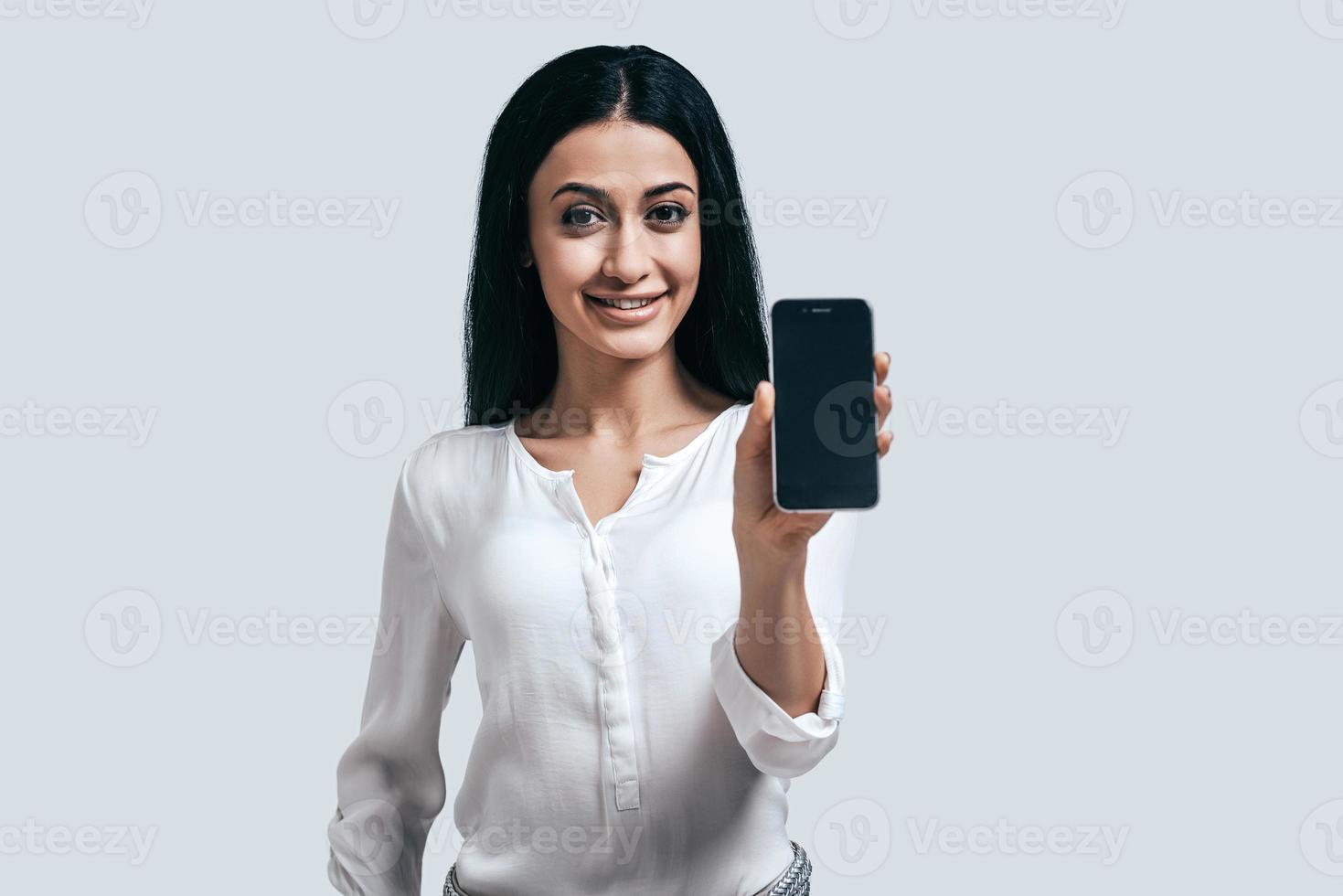 handig manier van verbinding. aantrekkelijk jong vrouw in wit overhemd Holding smartphone in full-front positie terwijl staand tegen grijs achtergrond foto