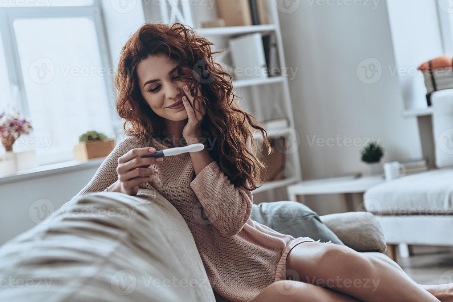 Tenslotte zwanger. aantrekkelijk jong Dames op zoek Bij zwangerschap test en glimlachen terwijl zittend Aan de sofa Bij huis foto