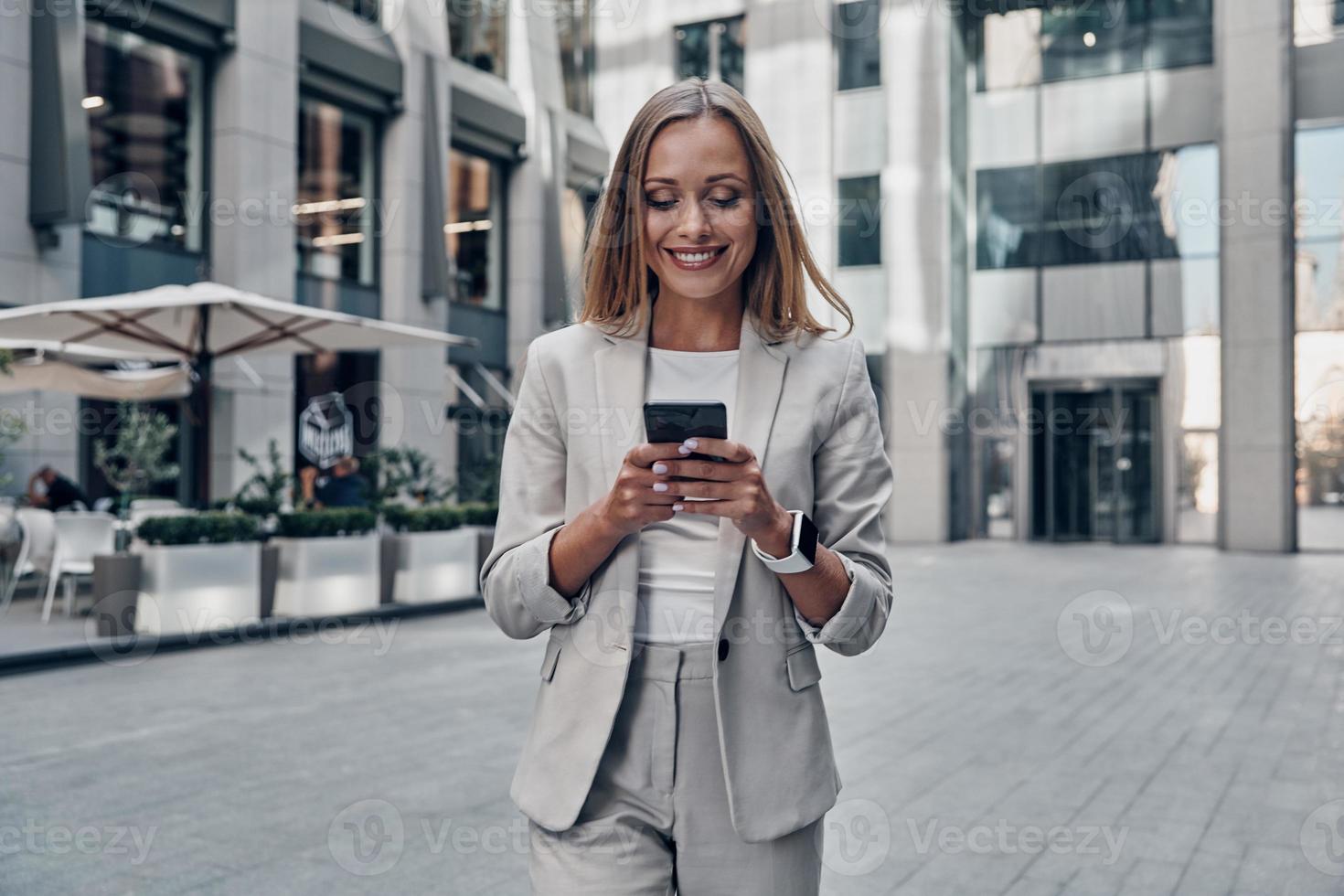 babbelen met cliënt. mooi jong vrouw in pak gebruik makend van slim telefoon en glimlachen terwijl staand buitenshuis foto