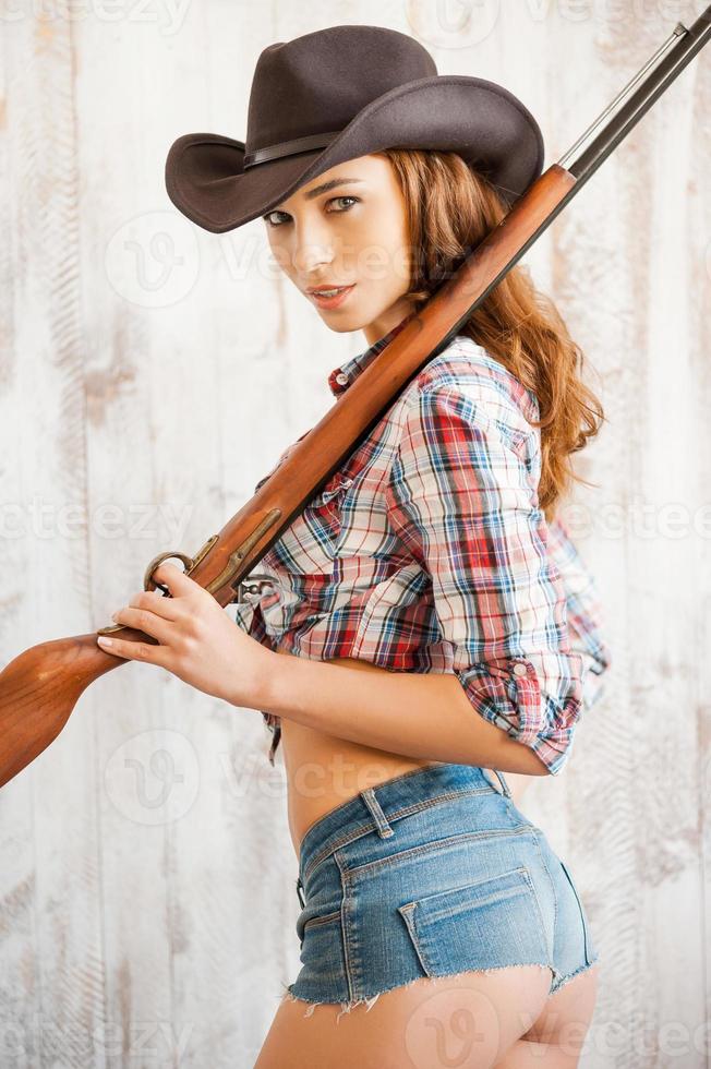 gevaarlijk schoonheid. mooi jong veedrijfster draag- geweer Aan haar schouders terwijl staand tegen de houten achtergrond foto