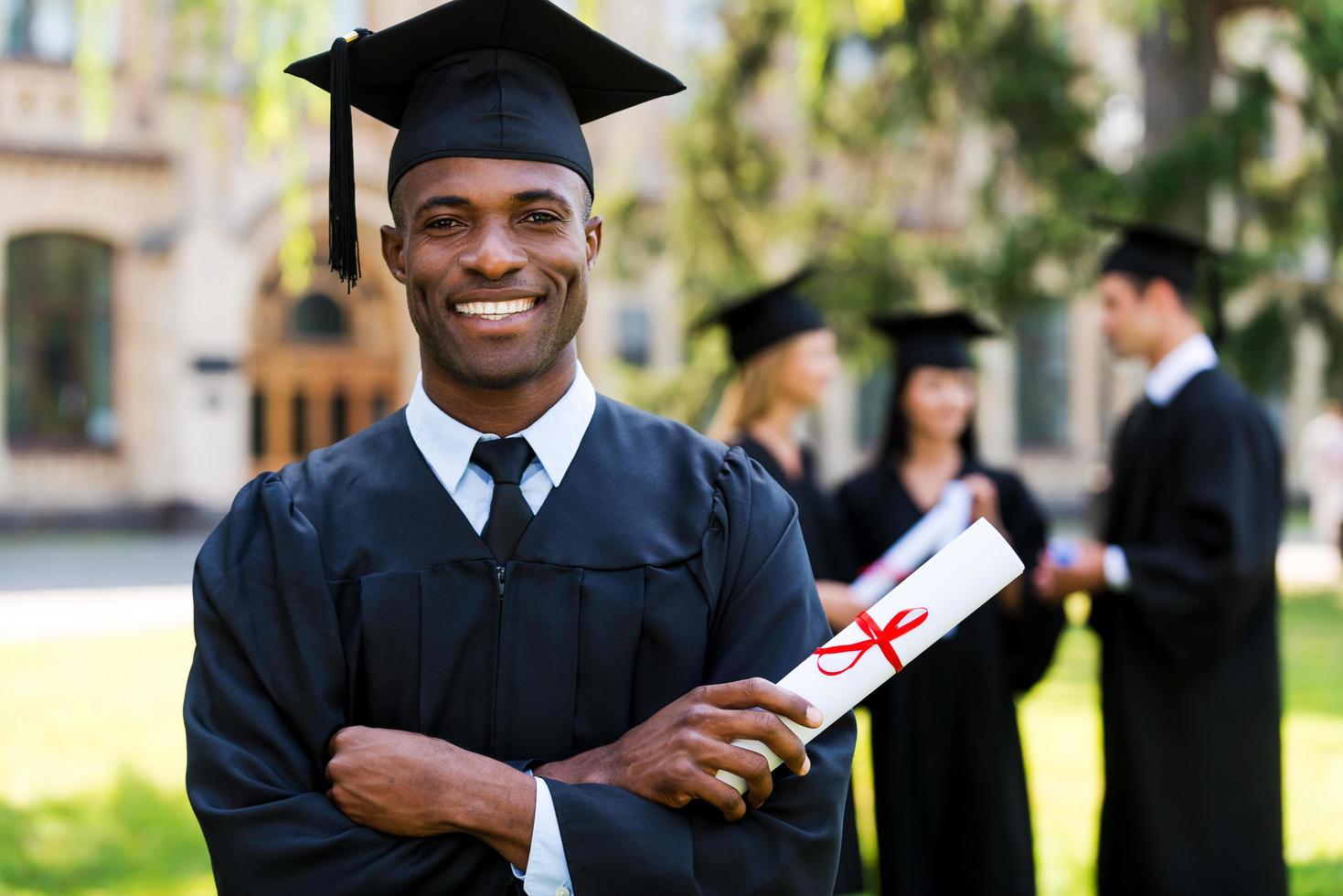 gelukkig afstuderen. gelukkig Afrikaanse Mens in diploma uitreiking jurken Holding diploma en glimlachen terwijl zijn vrienden staand in de achtergrond foto