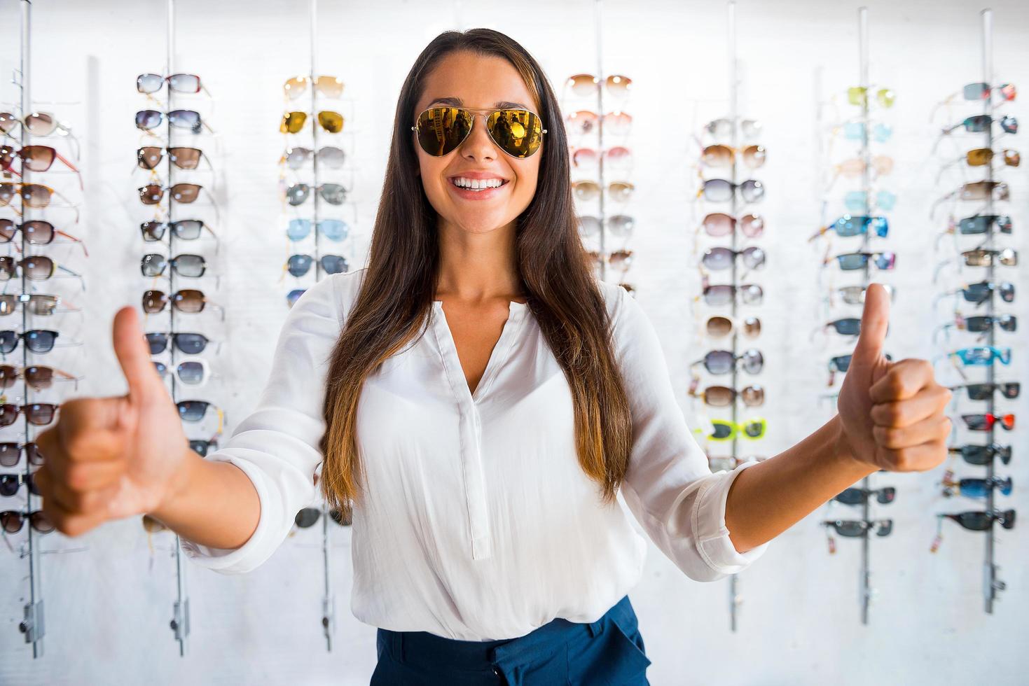 mooi zo keuze. mooi jong vrouw in zonnebril tonen haar duimen omhoog en glimlachen terwijl staand in optiek op te slaan foto