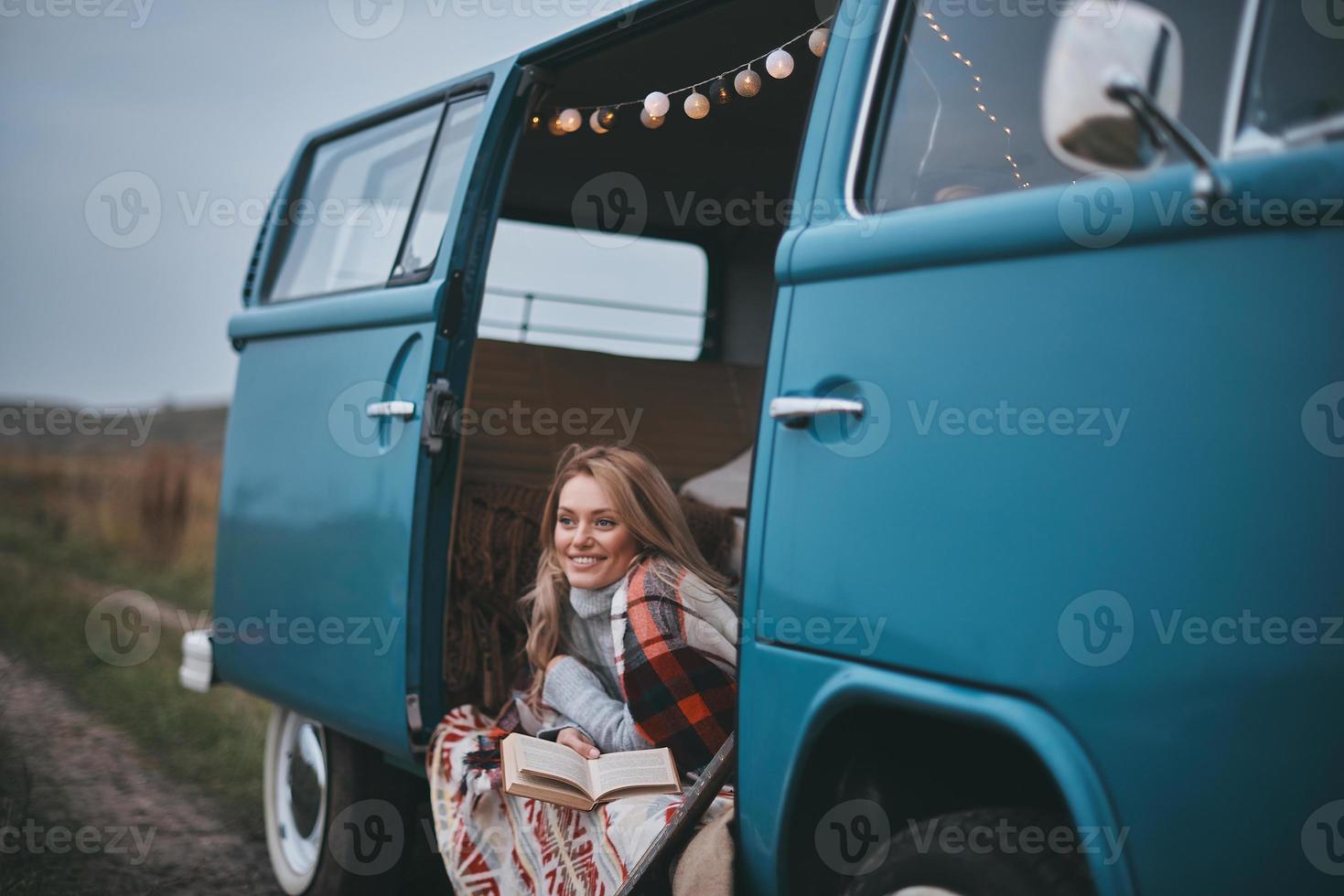 dromen over... aantrekkelijk jong vrouw gedekt met deken op zoek weg en glimlachen terwijl zittend binnen van de blauw retro stijl mini busje foto
