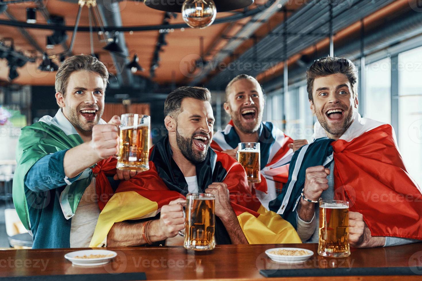 gelukkig jong mannen gedekt in Internationale vlaggen drinken bier en aan het kijken sport spel terwijl zittend in de kroeg foto