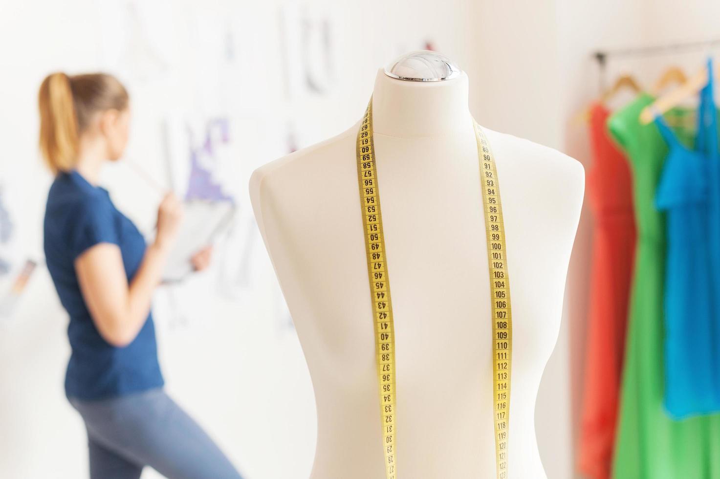 in zoeken van inspiratie. vrouw mode ontwerper op zoek Bij de schetsen hangende Aan de muur terwijl mannequin met meten plakband staand Aan voorgrond foto