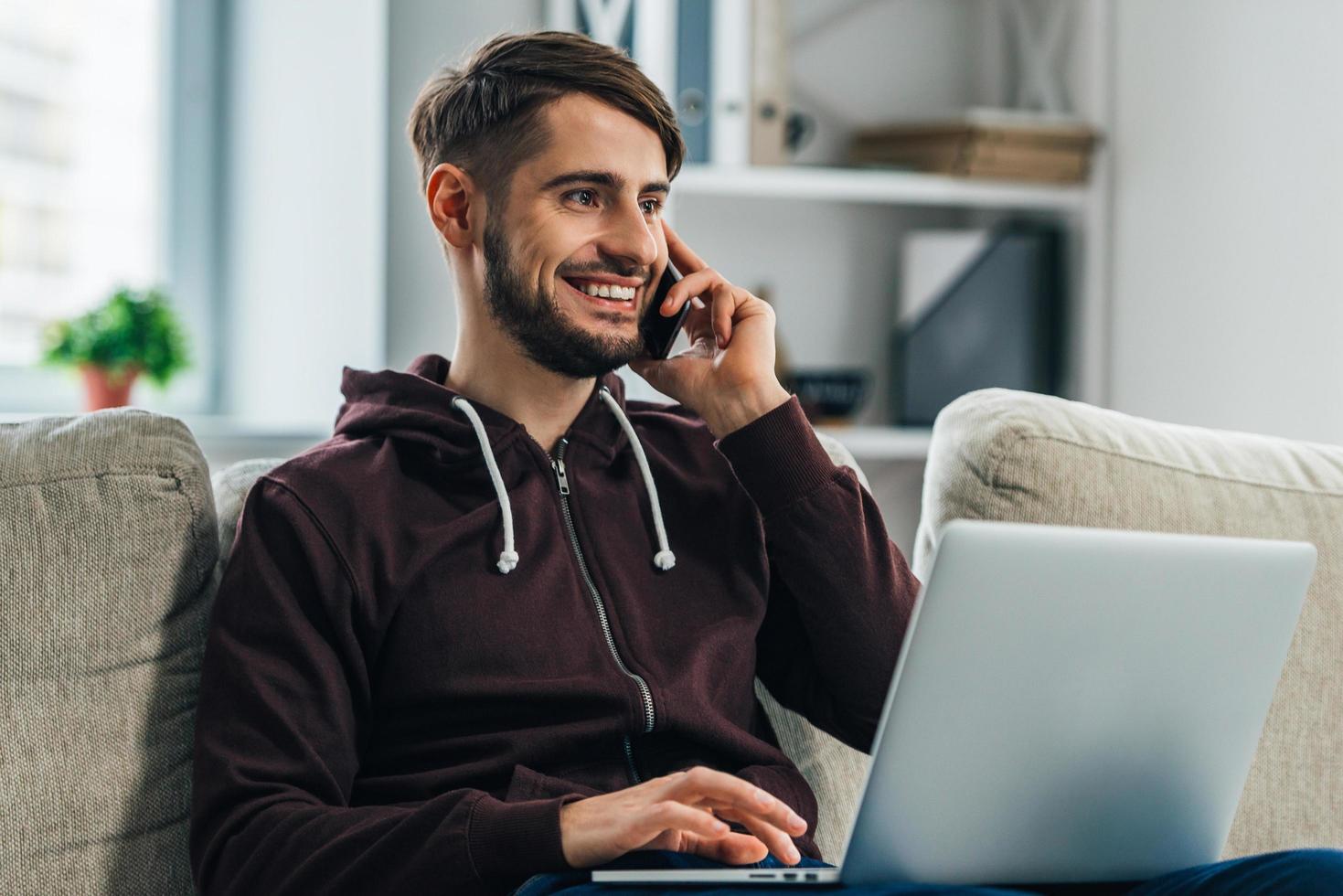 werken van huis met genoegen. vrolijk jong Mens gebruik makend van zijn laptop en pratend Aan mobiel telefoon terwijl zittend Aan bankstel Bij huis foto