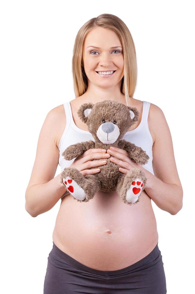 ervan uitgaand baby. gelukkig zwanger vrouw Holding teddy beer en glimlachen terwijl staand geïsoleerd Aan wit foto