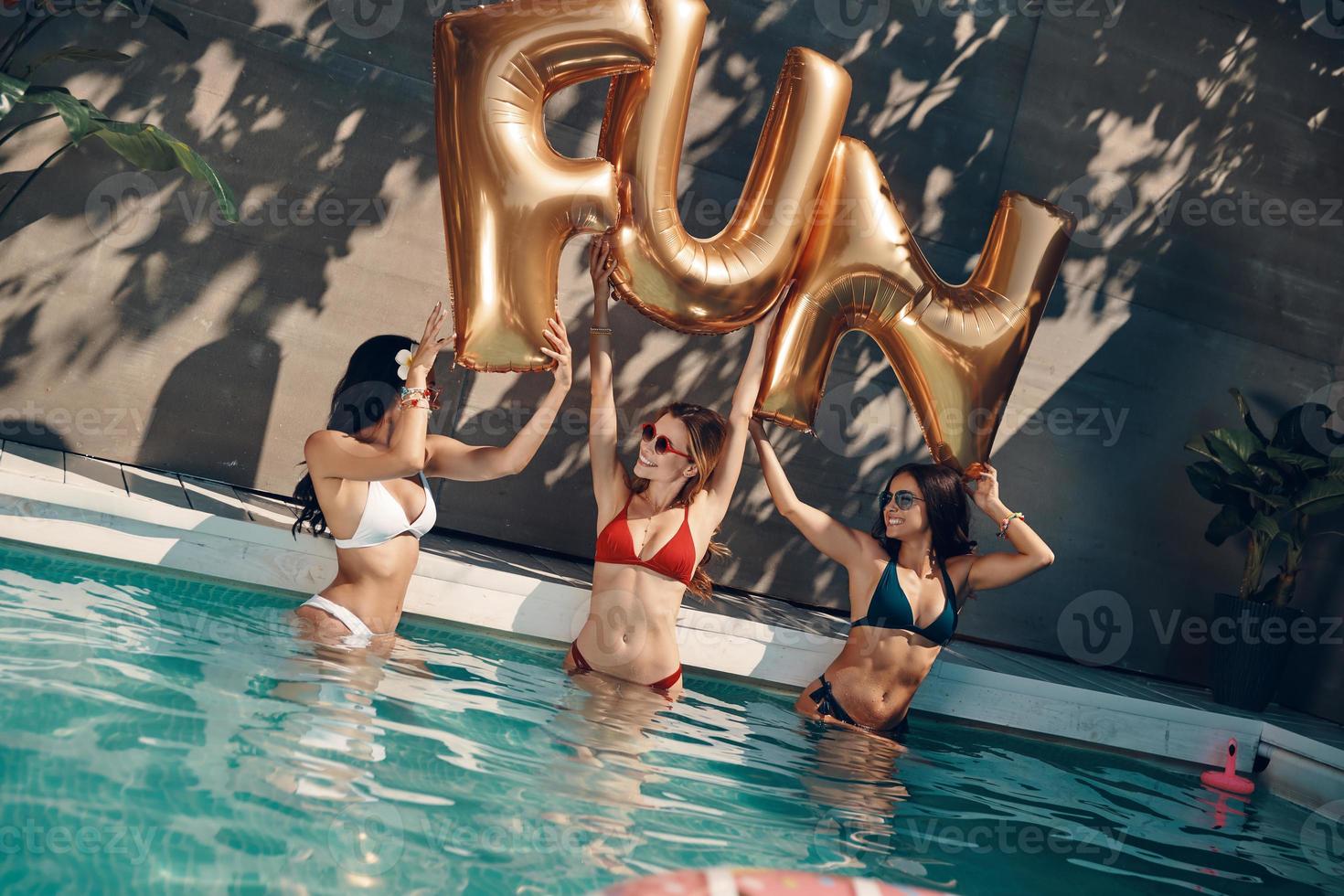 aantrekkelijk jong Dames in badmode glimlachen en hijs- omhoog ballonnen terwijl staand in de zwembad buitenshuis foto