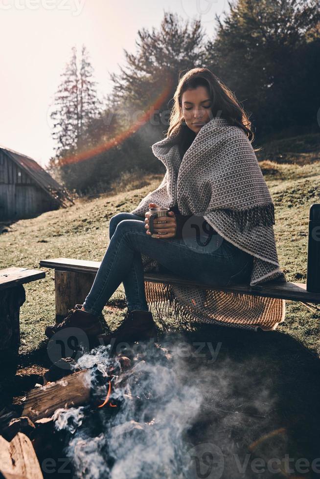 opwarming omhoog. mooi jong vrouw gedekt met deken Holding een mok terwijl zittend Aan de bank door de kampvuur foto