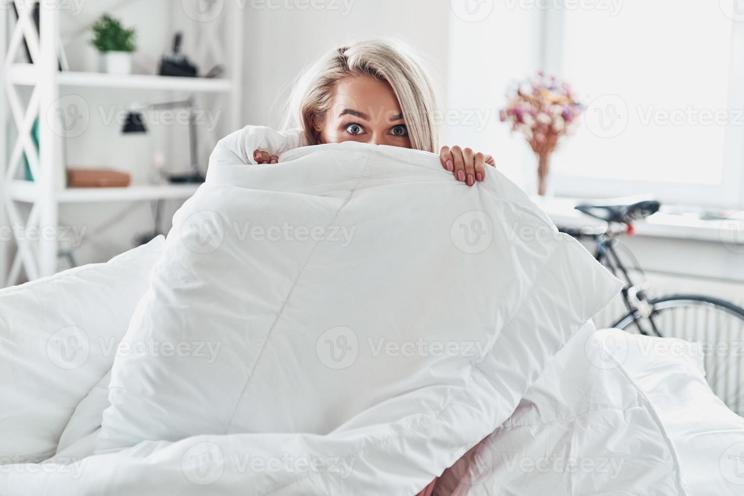 maandag ochtend. aantrekkelijk jong vrouw aan het bedekken voor de helft van haar gezicht met deken en op zoek Bij camera terwijl zittend in bed Bij huis foto