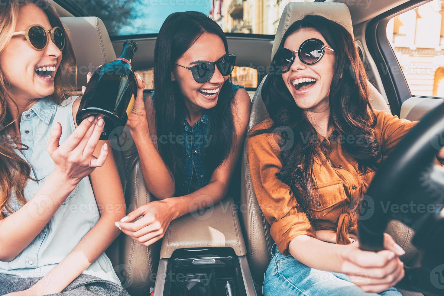 vieren een Super goed dag. drie mooi jong vrolijk Dames op zoek Bij elk andere met glimlach en Holding Champagne fles terwijl zittend in auto foto