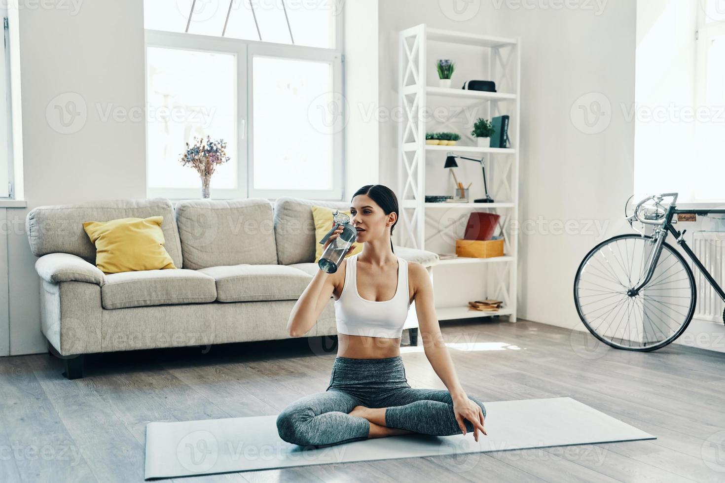 hydraterend. mooi jong vrouw in sport- kleding beoefenen yoga en drinken yoga terwijl uitgeven tijd Bij huis foto