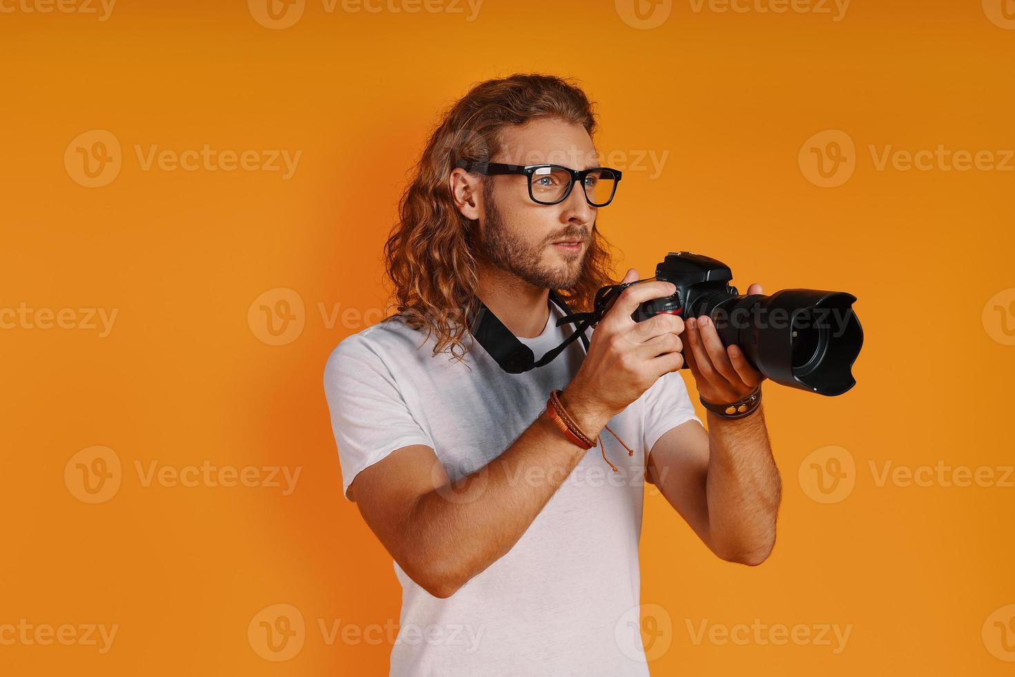 knap jong Mens in gewoontjes kleding gebruik makend van fotografisch camera terwijl staand tegen geel achtergrond foto