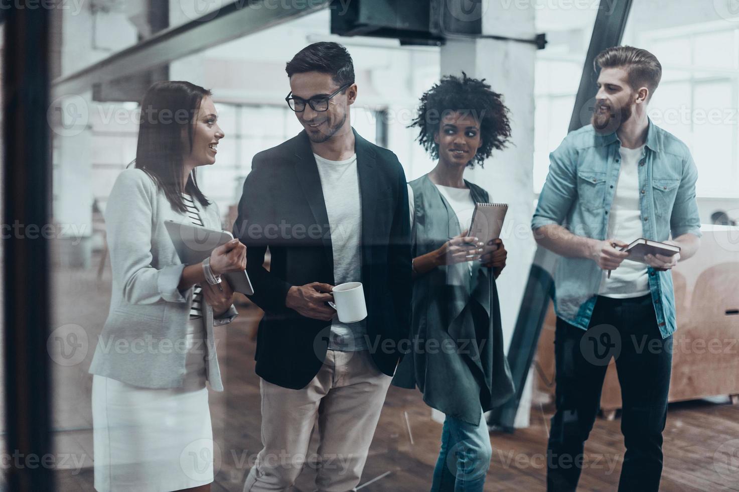 werken verder de kostgangers van een bureau. groep van jong mensen in slim gewoontjes slijtage pratend en glimlachen terwijl wandelen samen achter de glas muur in de creatief kantoor foto