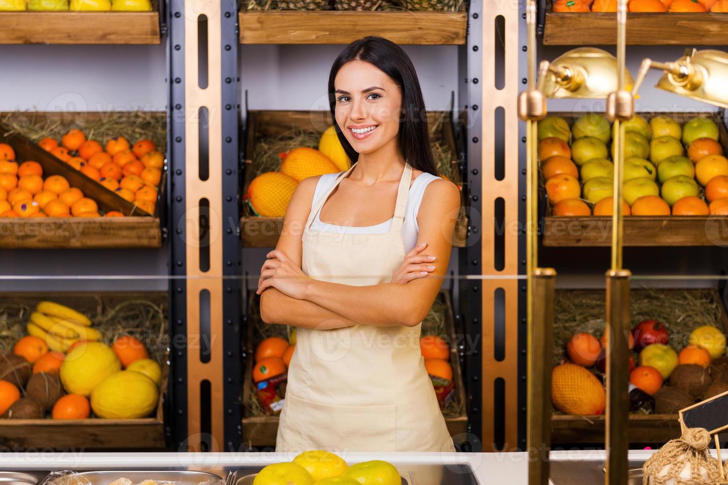 enkel en alleen de meest verse fruit in onze op te slaan. mooi jong vrouw in schort houden armen gekruiste en glimlachen terwijl staand in kruidenier op te slaan met verscheidenheid van fruit in de achtergrond foto