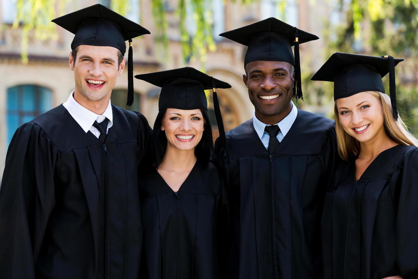 klaar naar helder toekomst. vier college afgestudeerden in diploma uitreiking jurken staand dichtbij naar elk andere en glimlachen foto
