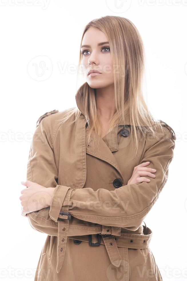 schoonheid in stijl. aantrekkelijk jong vrouw in jas houden armen gekruiste en op zoek weg terwijl staand tegen wit achtergrond foto